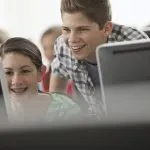 A school computer lab room, with rows of screens. Two young people looking intently at the screen.